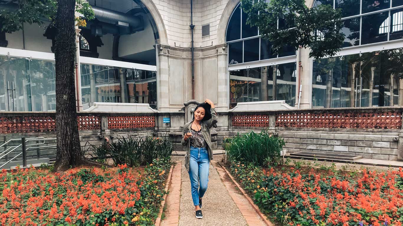 La imagen muestra a una mujer caminando por un jardín con flores rojas frente a un edificio histórico con arcos y paredes de vidrio. El entorno combina elementos arquitectónicos clásicos con áreas verdes, ofreciendo un espacio atractivo y tranquilo. El lugar parece ser el exterior del Museo Catavento, un centro cultural y educativo en São Paulo.