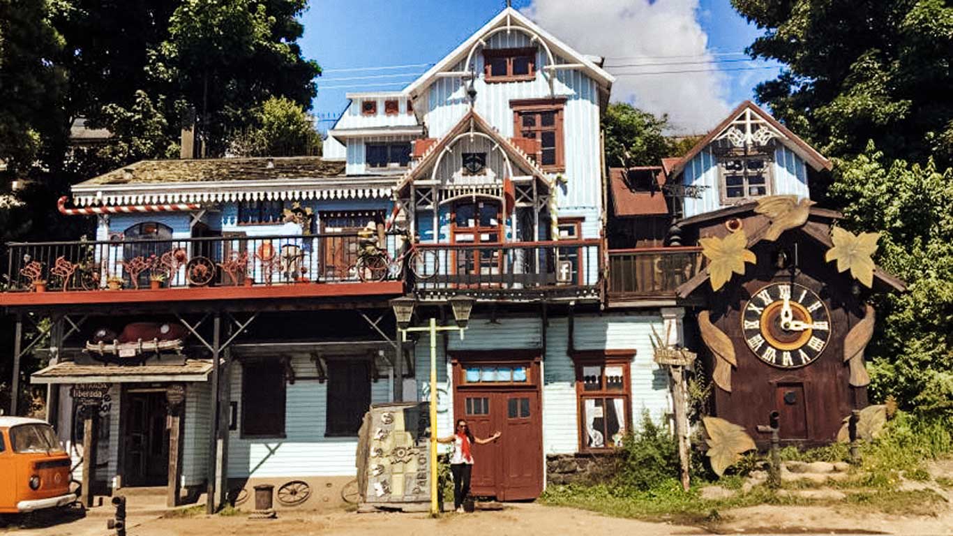 Imagen que muestra el Museo Pablo Fierro, una casa de estilo vintage con detalles arquitectónicos únicos en Puerto Varas. El edificio está decorado con elementos rústicos, como bicicletas antiguas, macetas y un gran reloj de madera en la fachada. Al frente, se observa una furgoneta clásica y una persona posando junto a la entrada, destacando la atmósfera artística y bohemia del lugar.