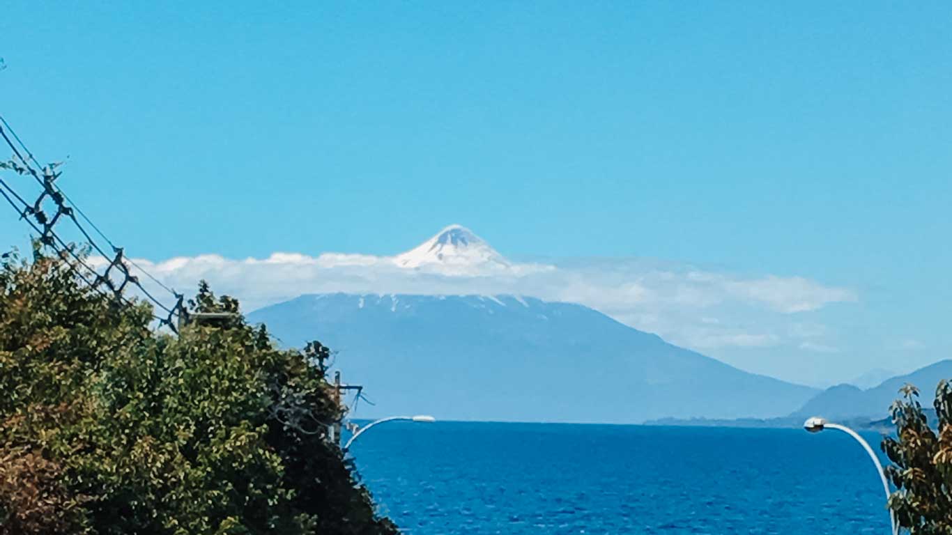 Imagen que muestra el volcán Osorno en la distancia, con su cima nevada sobresaliendo sobre un lago azul brillante bajo un cielo despejado. En el primer plano, se observan árboles y postes eléctricos que enmarcan la vista natural.