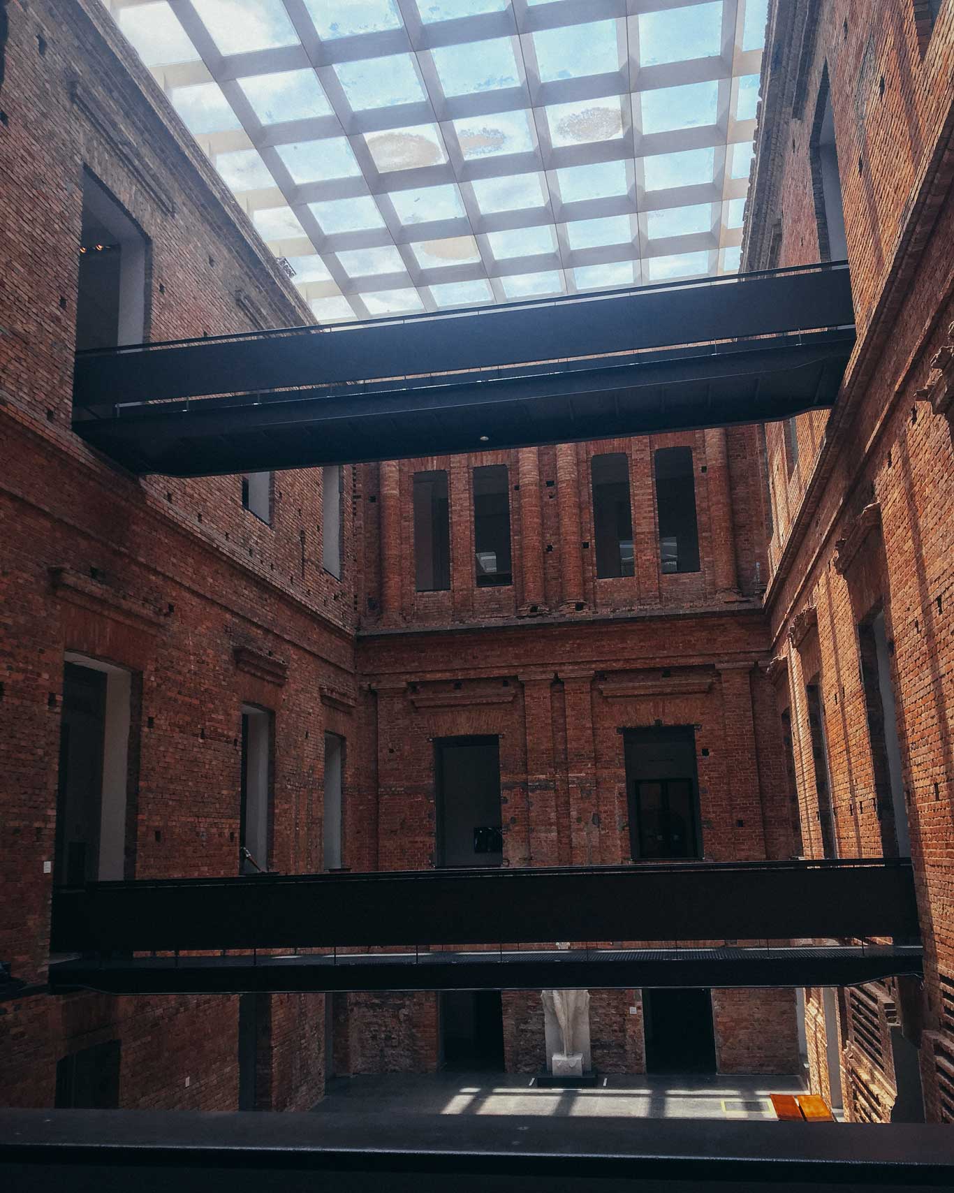 La imagen muestra el interior de un edificio histórico con paredes de ladrillo expuesto y un techo moderno con paneles de vidrio que permiten la entrada de luz natural. En el centro, hay pasarelas negras suspendidas que conectan diferentes áreas del espacio, típico de la Pinacoteca de São Paulo. La arquitectura combina elementos clásicos y contemporáneos, resaltando su carácter artístico y cultural.