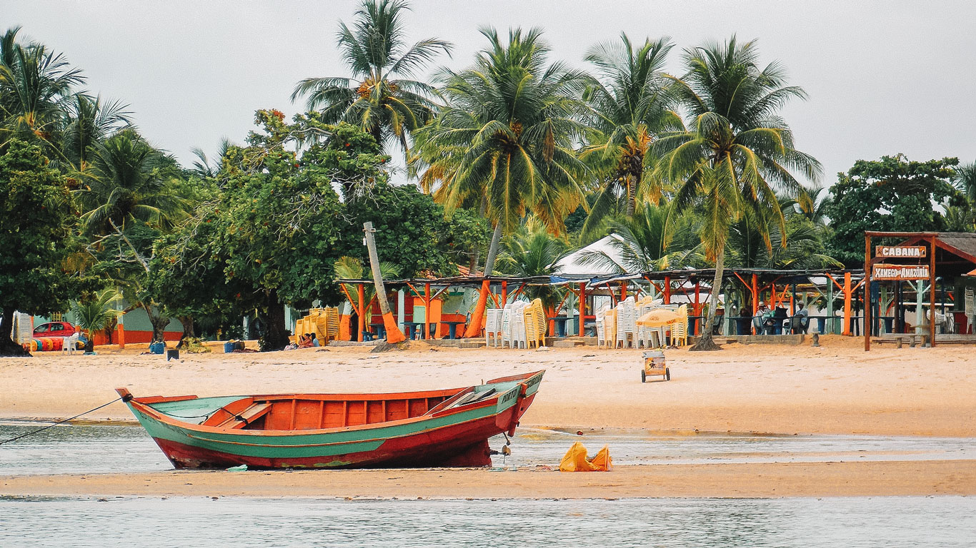 Playa de Coroa Vermelha en Porto Seguro, Brasil, con una colorida barca roja y verde reposando en la orilla. Al fondo, palmeras altas y frondosas se alinean junto a bares de playa con estructuras de madera y sillas apiladas. Un cartel que dice “Cabana Xamego da Amazônia” se encuentra a la derecha, completando esta escena tropical relajada.