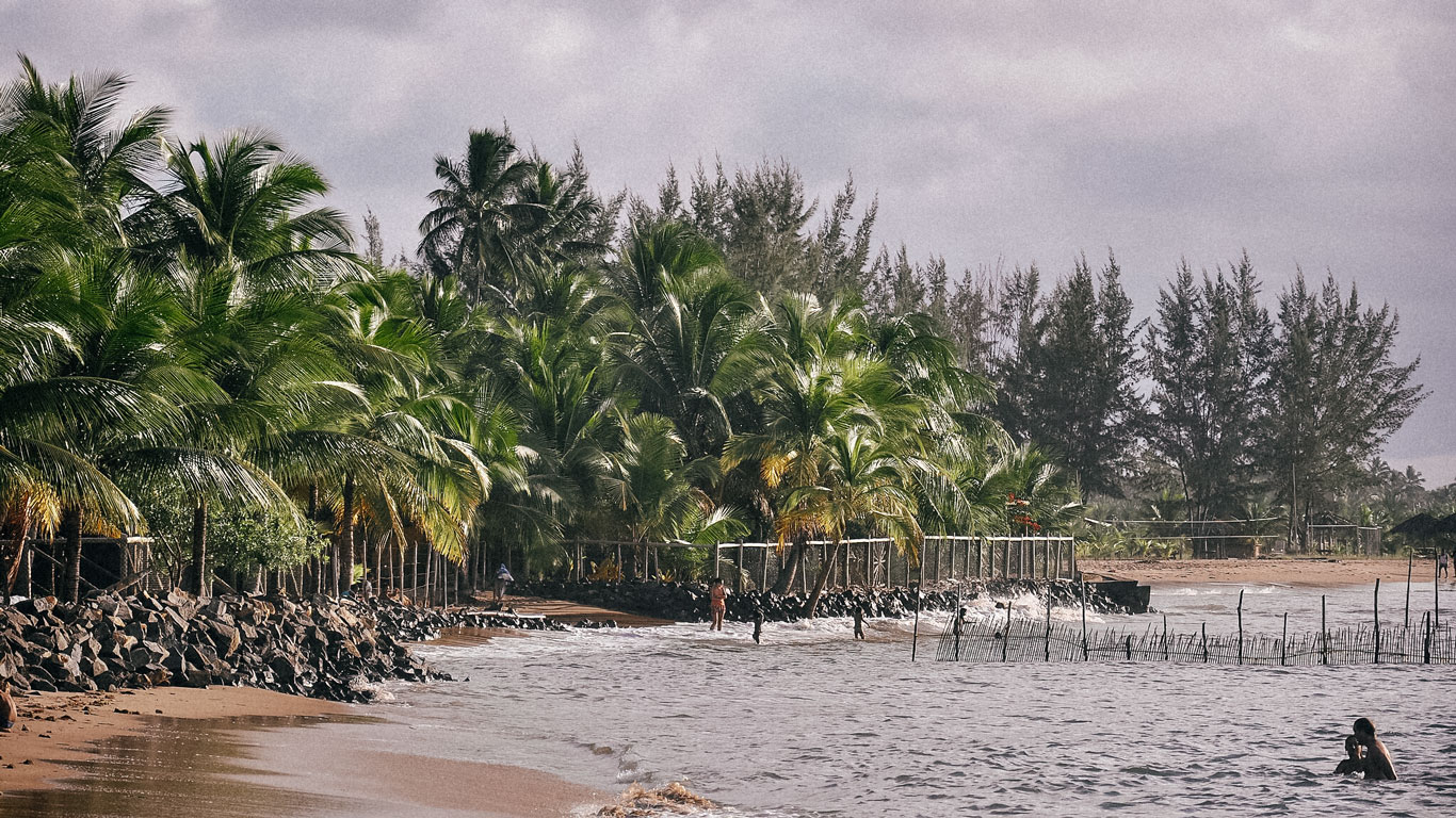 Playa de Mutá en Porto Seguro, Brasil, con aguas tranquilas y una franja de arena bordeada por palmeras verdes. Unas pocas personas disfrutan del mar, mientras una cerca improvisada hecha de palos de madera se extiende hacia el agua. La escena está enmarcada por un cielo nublado y rocas oscuras a lo largo de la costa.