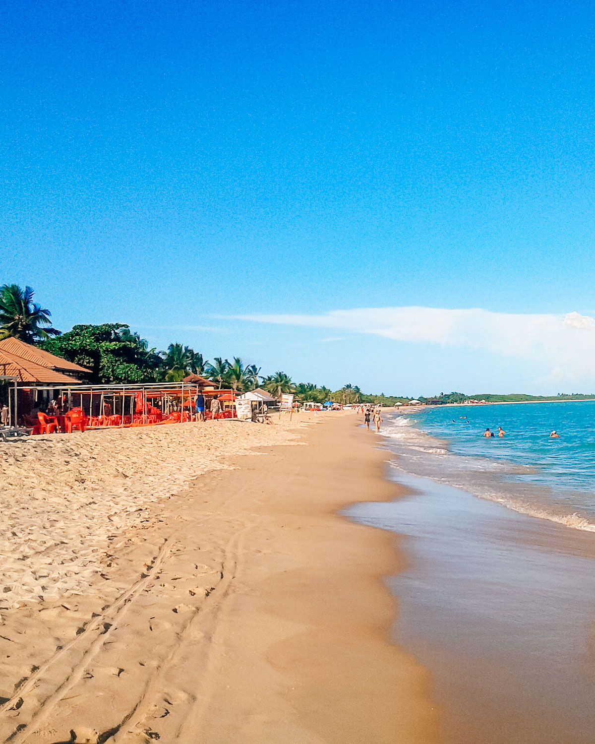 Playa de Taperapuã con una amplia franja de arena dorada y aguas tranquilas y azules, la mejor zona donde hospedarse en Poro Seguro. Varias personas disfrutan del mar, mientras que en la izquierda hay bares de playa con sillas y sombrillas rojas alineadas bajo estructuras de paja. El cielo despejado y las palmeras al fondo completan la escena de este popular destino turístico.