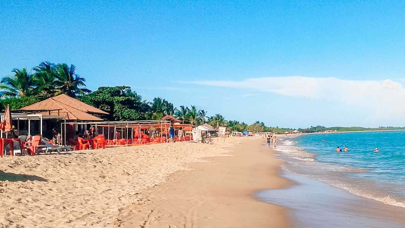 Playa de Taperapuã en Porto Seguro, Brasil, con una amplia franja de arena dorada y aguas tranquilas y azules. Varias personas disfrutan del mar, mientras que en la izquierda hay bares de playa con sillas y sombrillas rojas alineadas bajo estructuras de paja. El cielo despejado y las palmeras al fondo completan la escena de este popular destino turístico.