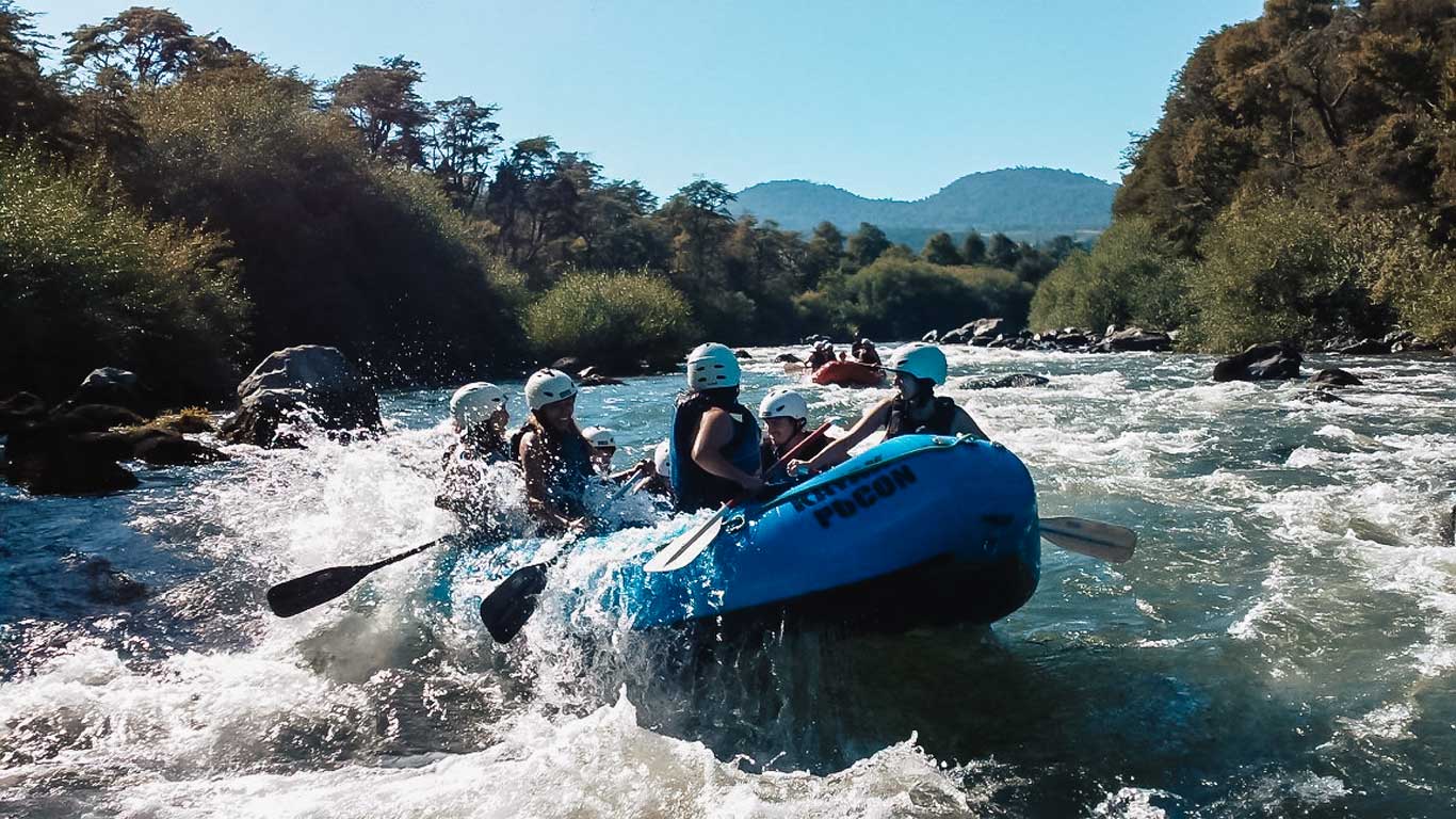 Imagen que muestra a un grupo de personas practicando rafting en un río de aguas rápidas en Pucón, Chile. Equipados con cascos y chalecos salvavidas, navegan en una balsa azul mientras atraviesan corrientes rodeadas de vegetación densa y montañas al fondo. Este tipo de actividad es ideal para los amantes de la aventura y la naturaleza, una experiencia emocionante en un entorno espectacular.