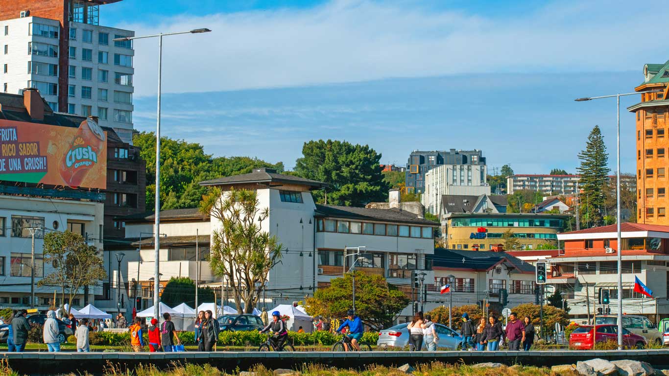 Imagen que muestra una escena urbana en Puerto Montt, Chile, con edificios modernos y tradicionales mezclados, rodeados de vegetación. En el primer plano, personas caminan y andan en bicicleta mientras disfrutan del espacio público bajo un cielo azul. El ambiente refleja el dinamismo de la ciudad con toques de naturaleza y modernidad en armonía.