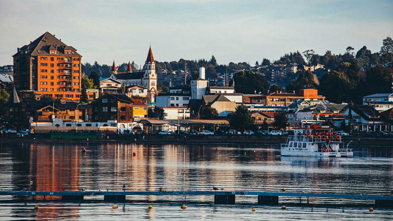 Imagen del centro de Puerto Varas, Chile, destacando su arquitectura típica con edificios de madera y la icónica iglesia del Sagrado Corazón de Jesús en el fondo. En el lago, un barco turístico con la inscripción "Puerto Varas" navega mientras los reflejos del atardecer iluminan el agua.