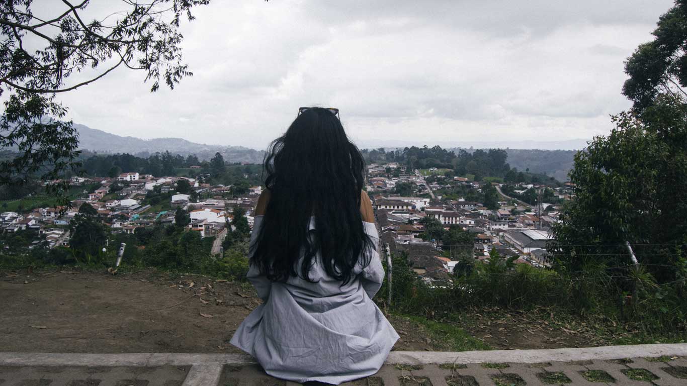 Una mujer con cabello largo y oscuro, sentada en un mirador, observa un pueblo de Salento rodeado de colinas y vegetación. El cielo está nublado, creando una atmósfera tranquila, mientras las casas blancas y los tejados de colores se extienden a lo largo del paisaje. La escena transmite una sensación de serenidad y conexión con la naturaleza.