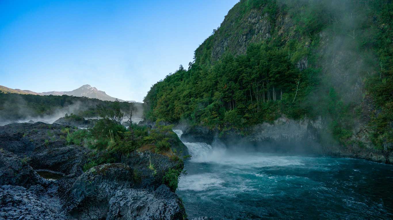 Imagen que muestra los Saltos de Petrohué, un impresionante paisaje natural en Chile, donde el agua cristalina fluye entre formaciones rocosas rodeadas de vegetación frondosa. En el fondo, se observa un pico montañoso bajo un cielo azul despejado, mientras una ligera neblina se levanta del agua, añadiendo un toque místico al lugar.