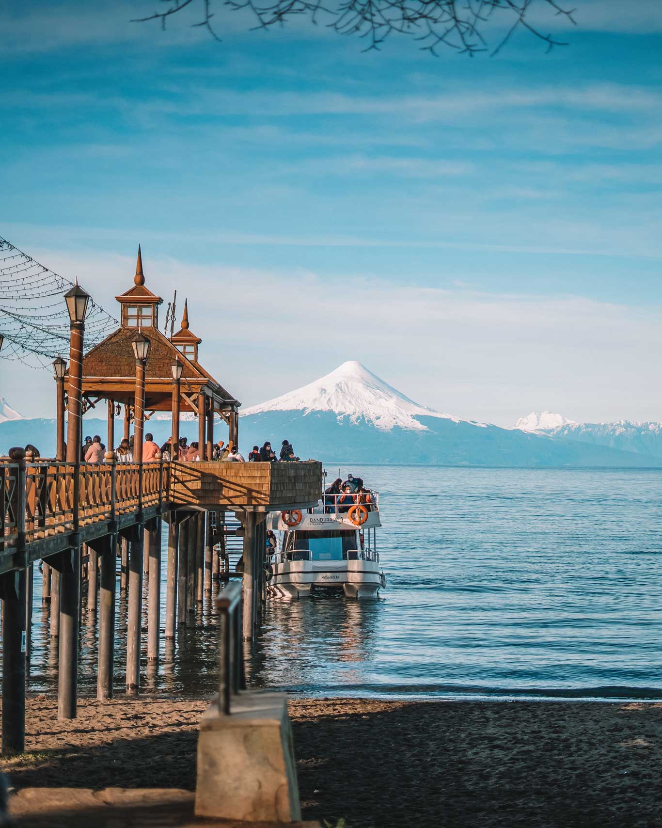 Imagen que muestra un muelle de madera en Puerto Varas, con un grupo de personas disfrutando de la vista al lago. En el agua, un barco turístico espera a los pasajeros, mientras al fondo se aprecia el imponente volcán Osorno cubierto de nieve, bajo un cielo azul claro. La escena combina arquitectura tradicional con un paisaje natural impresionante.