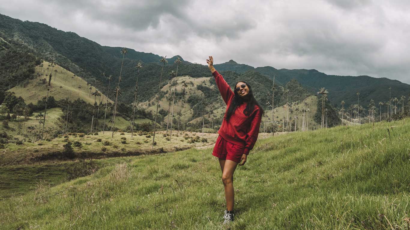 Una mujer vestida con un conjunto rojo sonríe y levanta una mano mientras posa en el Valle de Cocora, rodeada de verdes colinas y altas palmas de cera. Este lugar emblemático de Colombia es ideal para disfrutar de caminatas y vistas panorámicas y tiene excelentes alojamientos dónde alojarse en Salento.