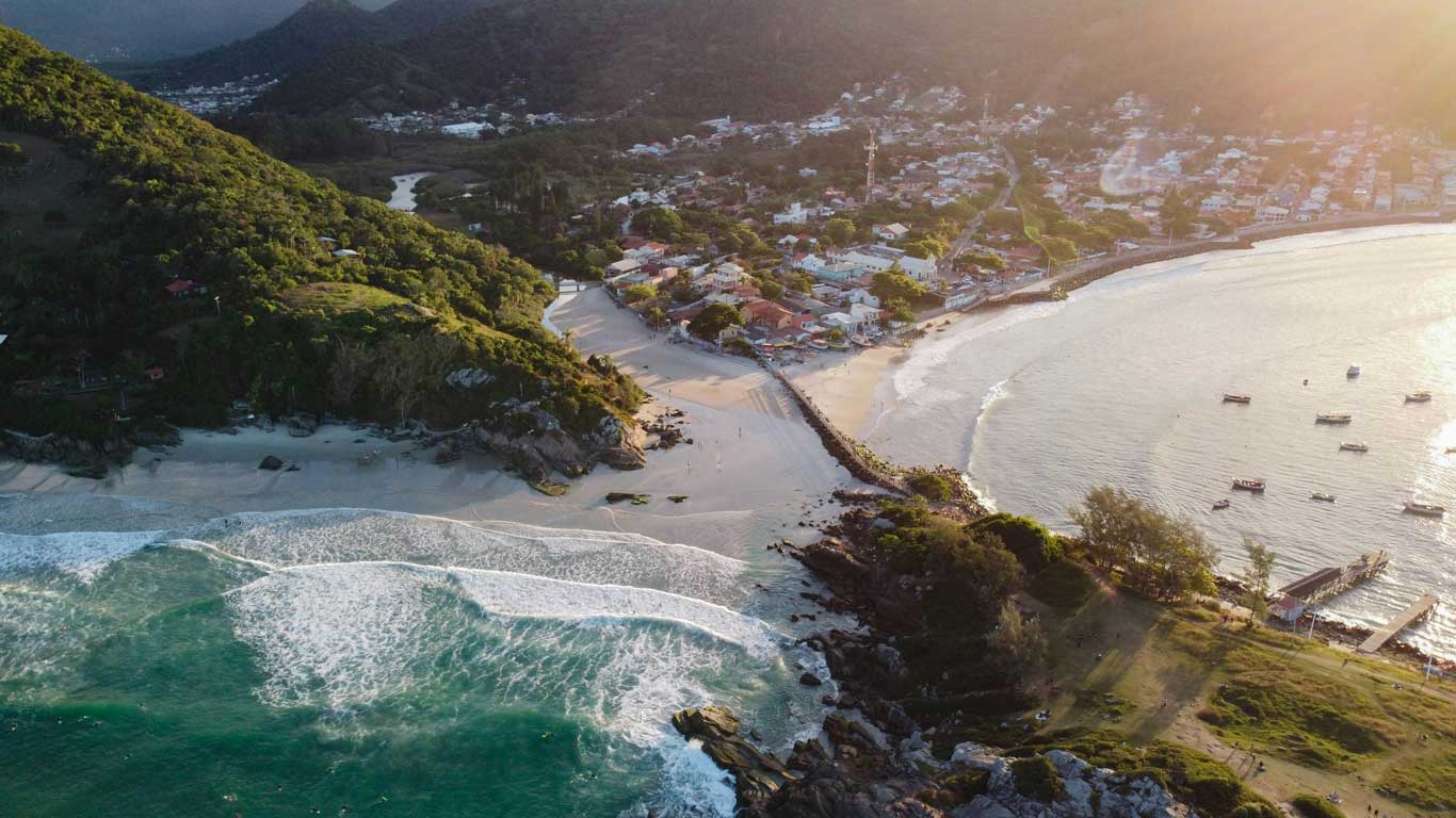 armacao-florianópolis Vista aérea de la playa Armação en Florianópolis, mostrando un paisaje que combina mar, arena y vegetación verde. Las olas rompen suavemente en la costa, mientras un pequeño pueblo con casas y calles pintorescas se extiende al fondo. La luz del atardecer ilumina el paisaje, resaltando su belleza natural y su ambiente tranquilo.
