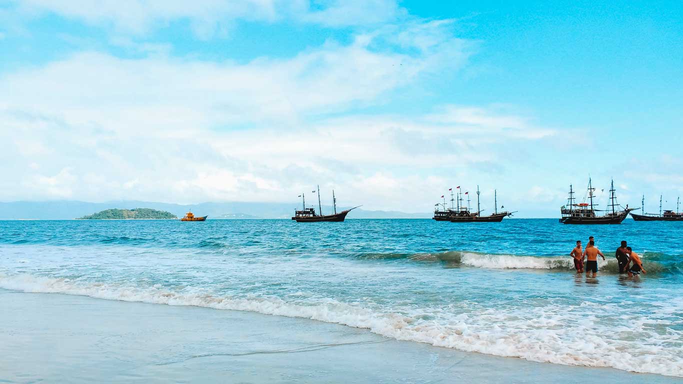 canasvieira-florianopolis Playa de aguas cristalinas en Canasvieiras, Florianópolis, con barcos estilo pirata navegando en el horizonte. Personas disfrutan del agua cerca de la orilla, mientras una pequeña isla verde se asoma en el fondo. El cielo azul y despejado complementa este ambiente tropical y relajante.