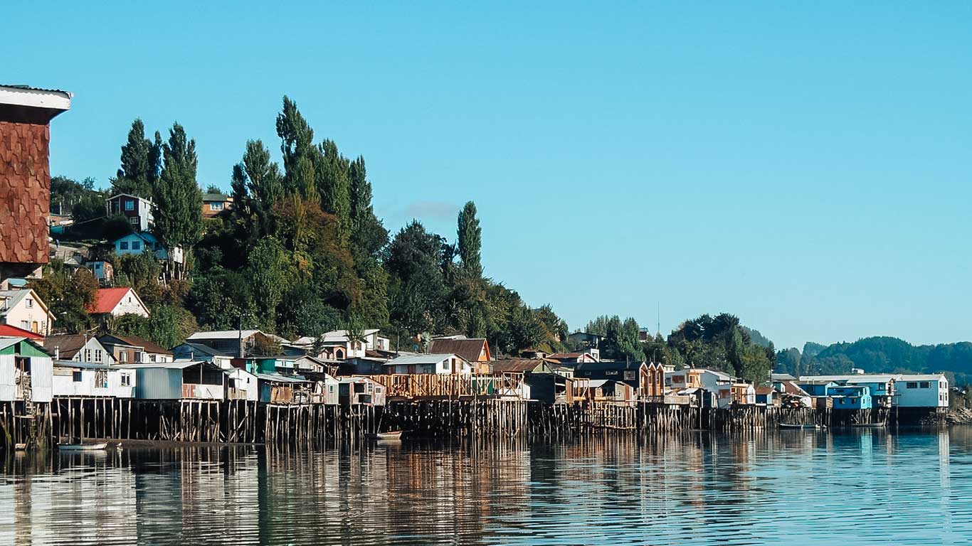 La imagen muestra los icónicos palafitos de Castro, Chiloé, un conjunto de coloridas casas de madera construidas sobre pilotes a la orilla del agua. El reflejo de las viviendas en el agua crea una escena pintoresca, mientras que el fondo está compuesto por colinas verdes con más casas dispersas entre los árboles. El cielo despejado y la luz natural resaltan los tonos vibrantes de la arquitectura típica de la isla.