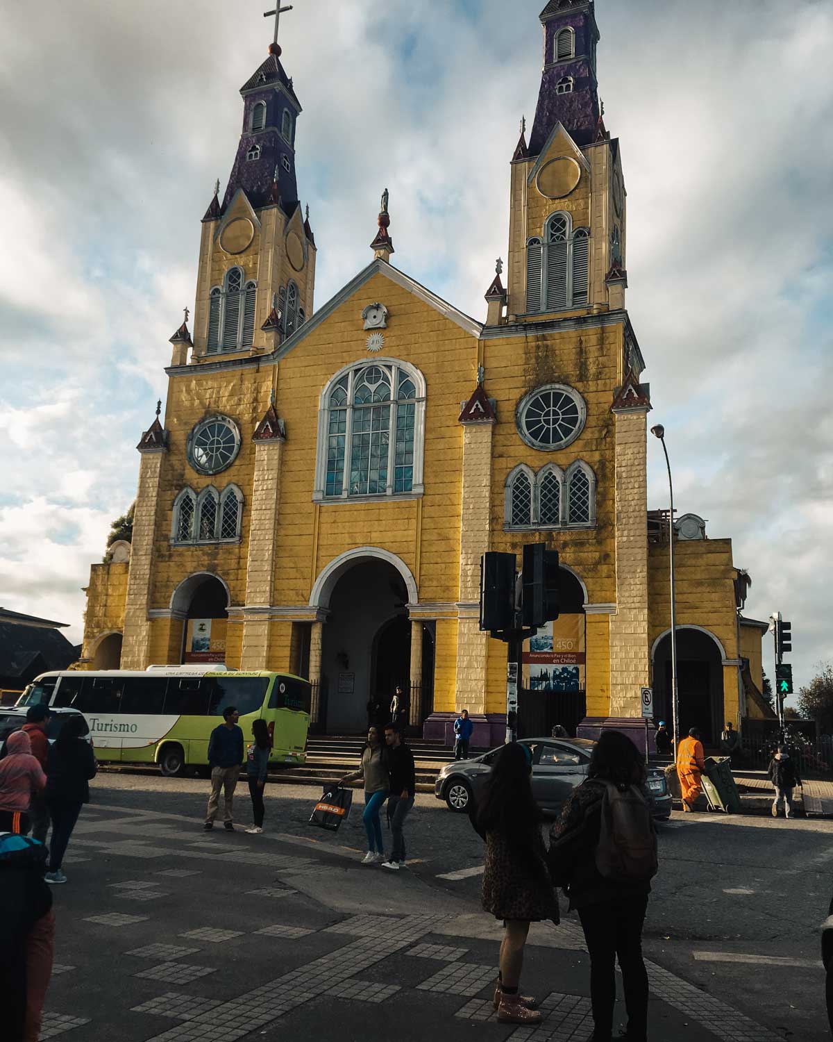 La imagen muestra la Iglesia de San Francisco en Castro, Chiloé, una de las iglesias patrimoniales de madera de la isla. Su fachada amarilla con detalles morados y sus dos altas torres la hacen destacar contra el cielo nublado. En el primer plano, varias personas caminan por la calle y la plaza, mientras un autobús de turismo y algunos autos están estacionados cerca, reflejando la actividad urbana de la zona.