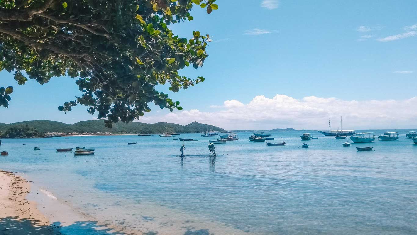 Playa en Búzios con aguas cristalinas y barcos pequeños anclados cerca de la orilla. Dos personas trabajan en el agua, probablemente pescando, mientras un gran velero destaca en el fondo. La escena está enmarcada por ramas de árboles, con colinas verdes al fondo y un cielo despejado.