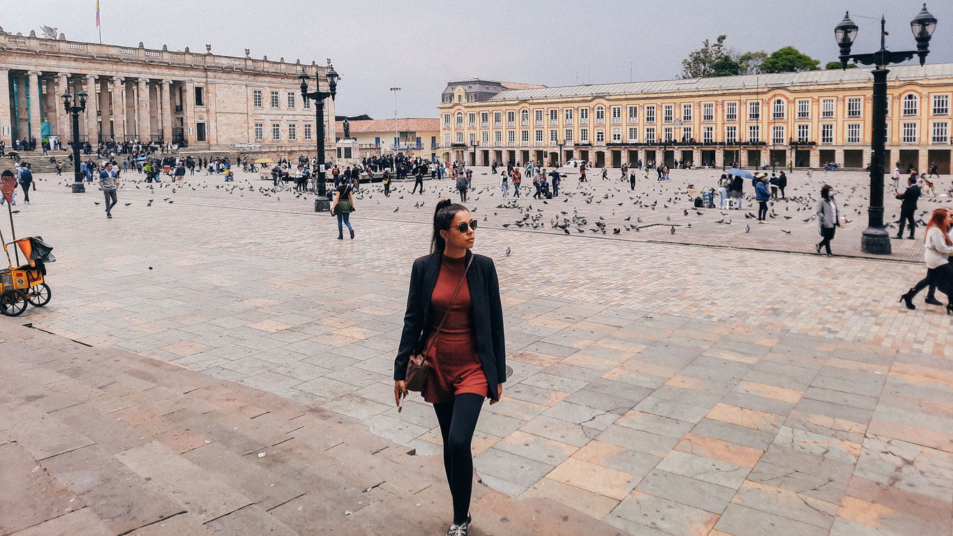 La imagen muestra una mujer caminando por una plaza amplia, rodeada de edificios históricos con arquitectura colonial en tonos beige y amarillo. En el fondo se aprecian columnas imponentes y una plaza llena de actividad, con personas y palomas. El ambiente sugiere un lugar turístico y cultural en Bogotá, Colombia.