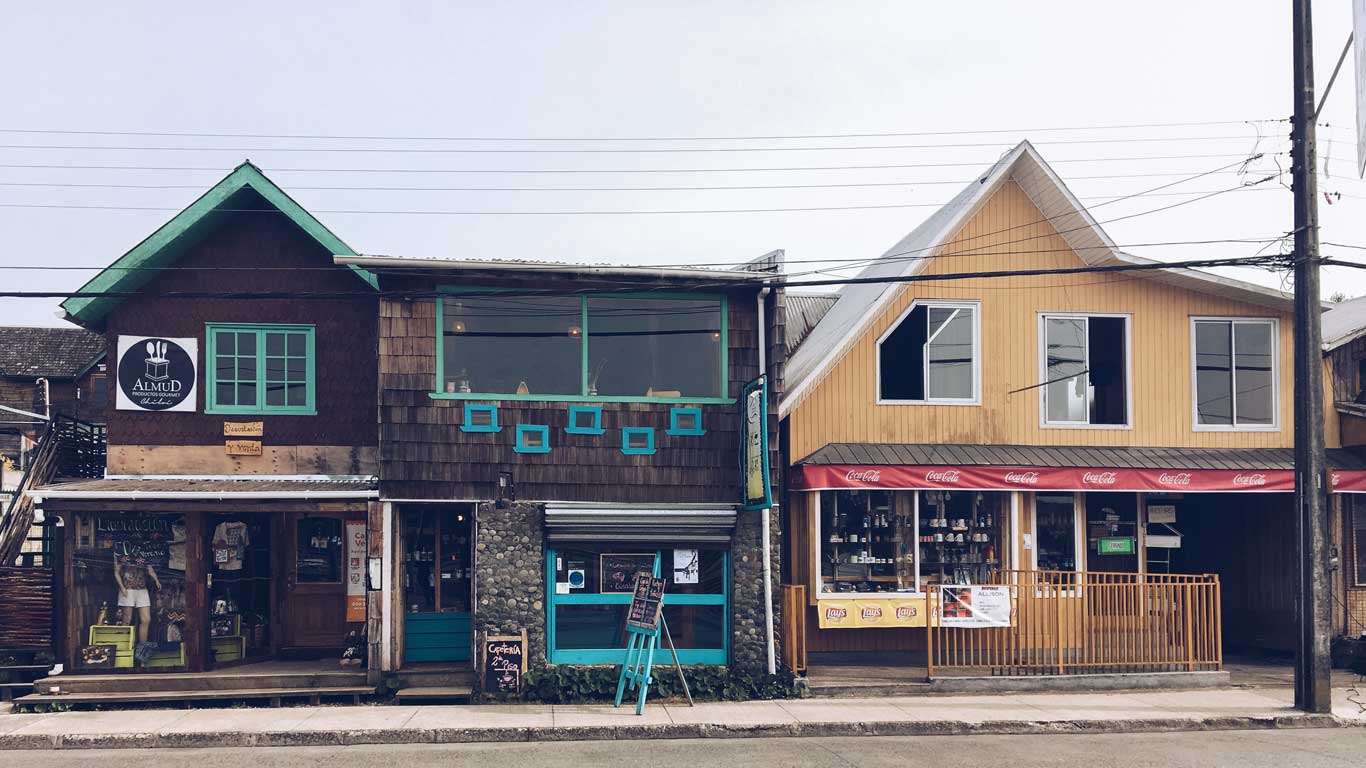 La imagen muestra dos edificios de madera con negocios en la planta baja en una calle de Chiloé, Chile. A la izquierda, hay una cafetería y tienda con un letrero que dice "ALMUD" y ventanas con marcos turquesas, mientras que a la derecha, hay un almacén con una fachada amarilla y un toldo rojo de "Coca-Cola". Los edificios tienen un estilo rústico, con techos inclinados y detalles artesanales, representando la arquitectura típica de la región.