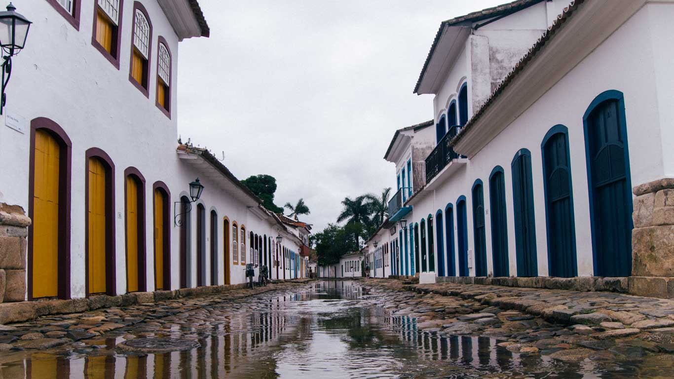 La imagen muestra una calle empedrada de Paraty, Brasil, con casas coloniales blancas de puertas y ventanas en tonos amarillos y azules. La calle está parcialmente inundada, reflejando las fachadas de los edificios y el cielo nublado, un fenómeno característico de la ciudad debido a las mareas. En el fondo, se observan palmeras y faroles coloniales, resaltando el encanto histórico y tropical de este destino.