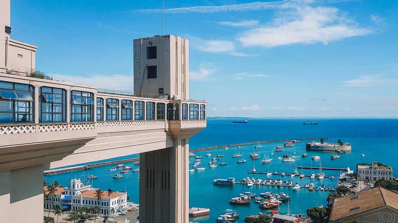 Vista del Elevador Lacerda en Salvador, Brasil, con su arquitectura icónica que conecta la Ciudad Alta con la Ciudad Baja. En el fondo, el puerto con numerosas embarcaciones y el océano azul se extienden bajo un cielo despejado. La imagen destaca el contraste entre la infraestructura histórica y el paisaje marítimo vibrante.