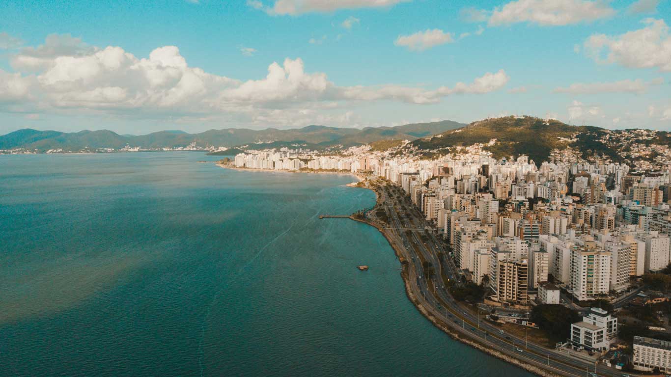 Vista aérea de la ciudad costera de Florianópolis, en Santa Catarina, Brasil. La imagen muestra una amplia bahía con agua azul y una línea de edificios altos a lo largo de la costa, con colinas verdes al fondo. El cielo está despejado con algunas nubes dispersas, resaltando la belleza natural y urbana del lugar.