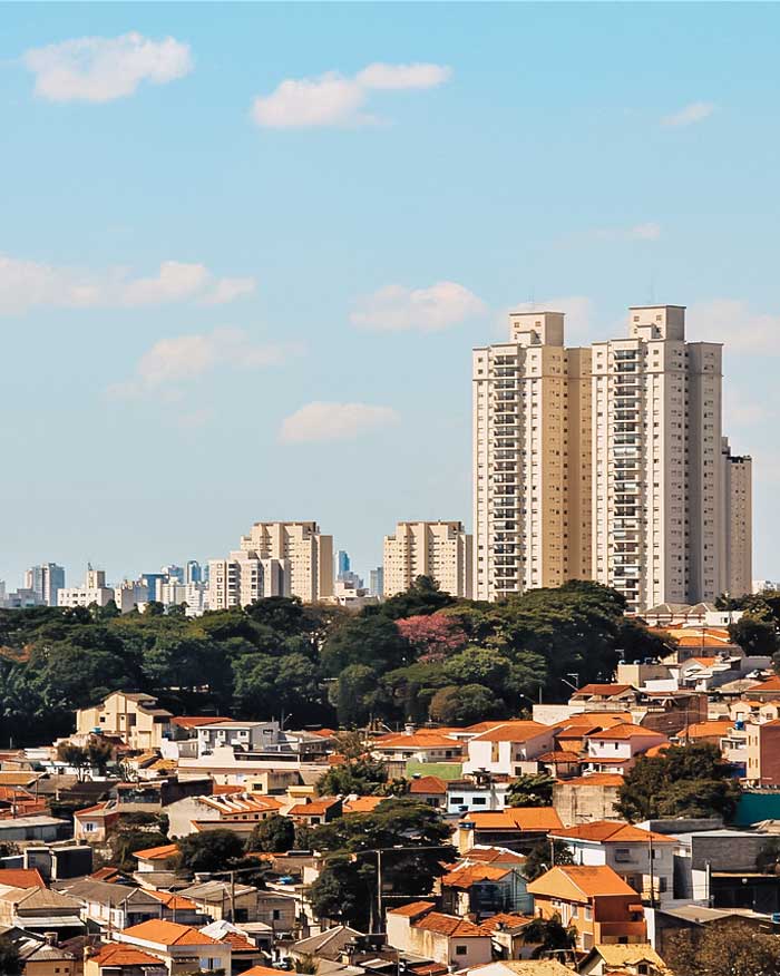 La imagen muestra una vista urbana de una zona residencial con casas de techos rojos en primer plano, rodeadas de vegetación, y edificios altos al fondo bajo un cielo azul despejado. El contraste entre las casas bajas y los rascacielos resalta la mezcla arquitectónica y el crecimiento urbano de la ciudad. Es un paisaje típico de una metrópoli en desarrollo, donde la naturaleza y la construcción conviven.