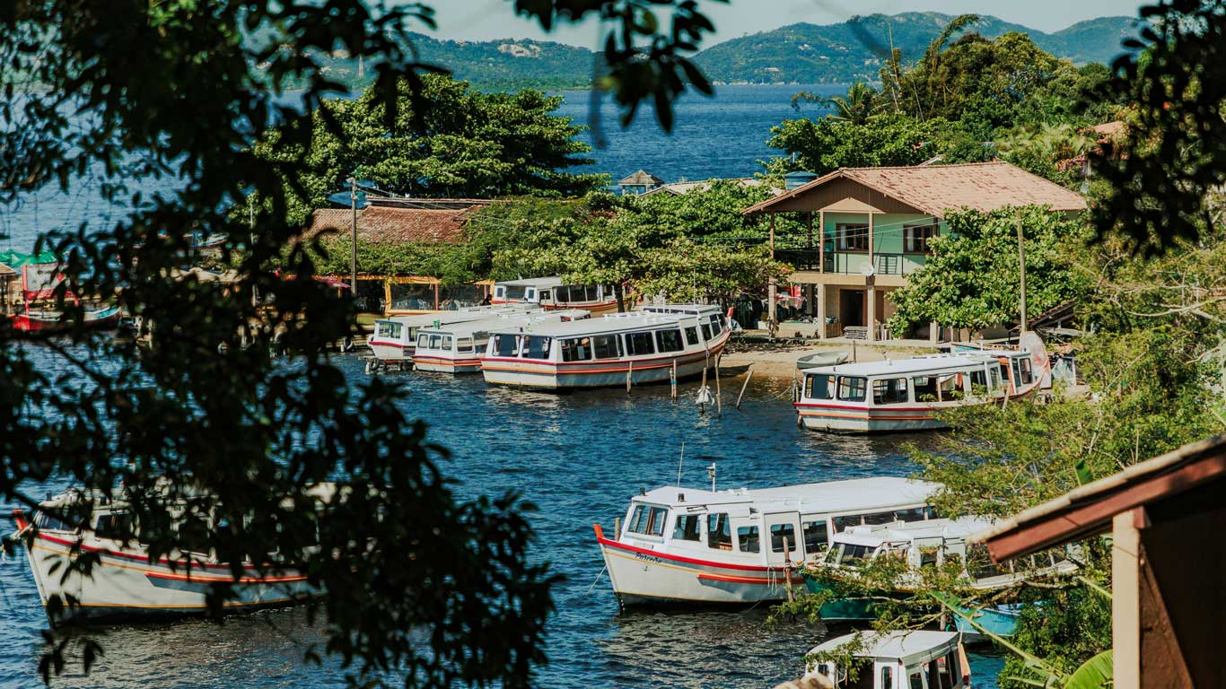 lagoa-da-conceicao-florianopolis Vista de una pintoresca zona junto a la Lagoa da Conceição en Florianópolis, Brasil, con barcos turísticos atracados en el agua y una casa rodeada de vegetación. Este lugar combina naturaleza y comodidad, ideal para quienes buscan dónde alojarse en Florianópolis cerca de la laguna.