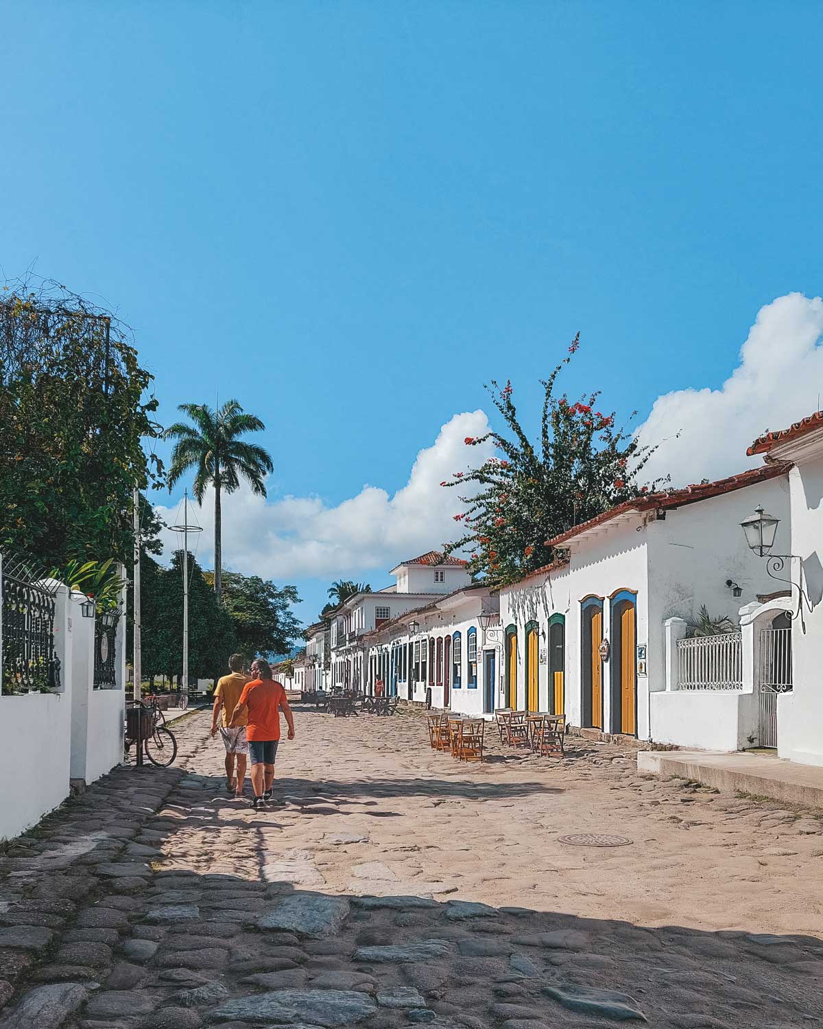 La imagen muestra una calle empedrada en Paraty, Brasil, con casas coloniales blancas de puertas y ventanas en colores vibrantes. Dos personas caminan de espaldas bajo un cielo azul despejado, rodeadas de mesas al aire libre y detalles florales en las fachadas. Al fondo, palmeras y vegetación complementan la escena tranquila y pintoresca de este destino histórico.