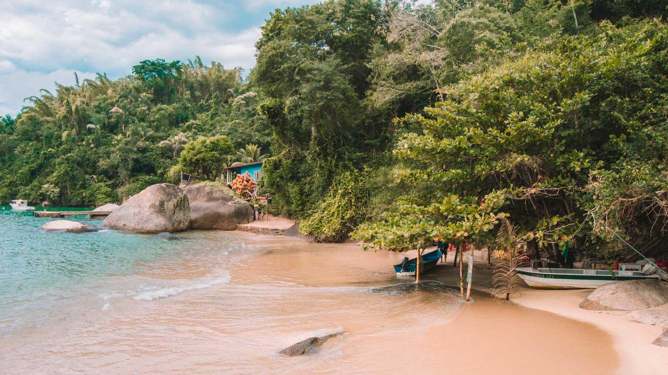 La imagen muestra una playa escondida en Paraty, Río de Janeiro, con arena dorada, aguas cristalinas y grandes rocas junto a la orilla. La densa vegetación tropical rodea el lugar, con árboles y arbustos extendiéndose hasta la playa, creando un ambiente selvático. Al fondo, una pequeña casa azul y varias embarcaciones resaltan la conexión entre la comunidad local y el mar, en un rincón tranquilo y paradisíaco.