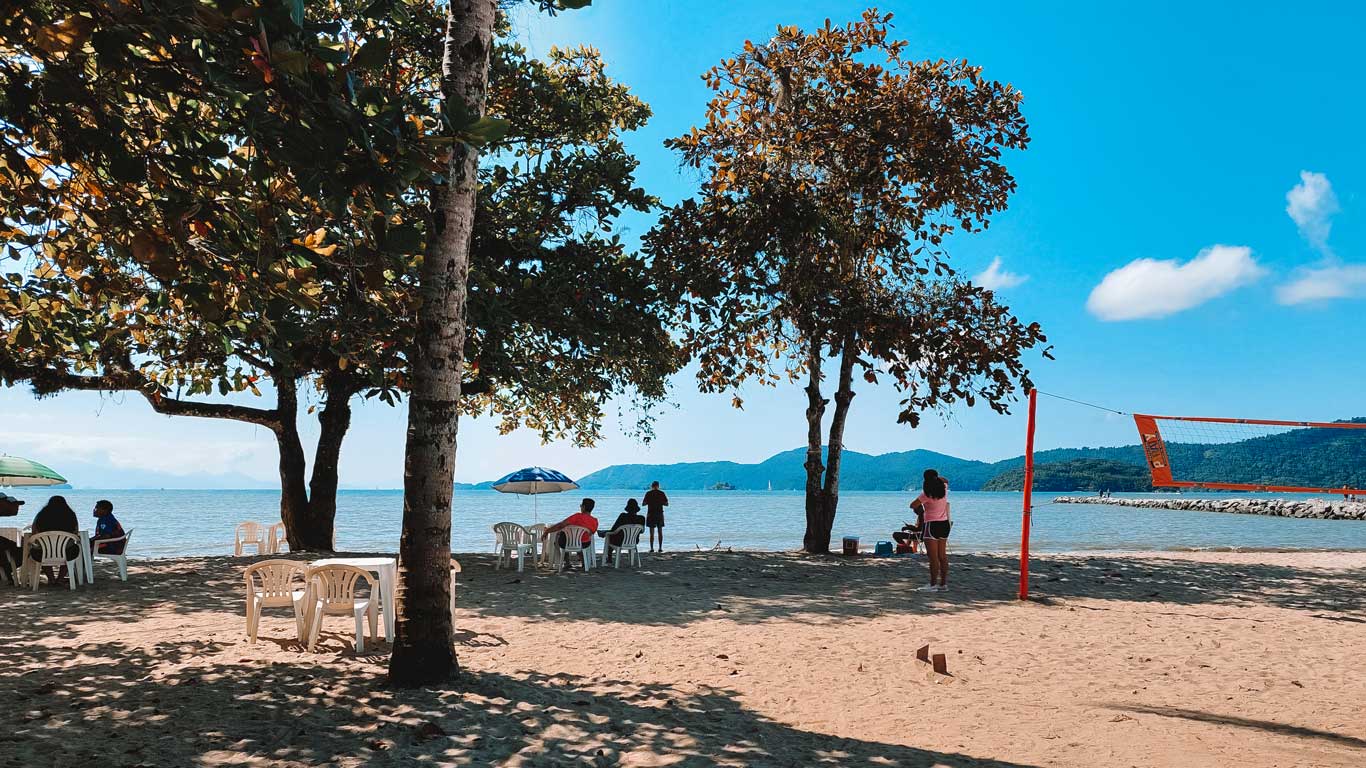 La imagen muestra la Playa de Pontal en Paraty, Brasil, con arena dorada, árboles que dan sombra y mesas blancas bajo sombrillas. Algunas personas están sentadas disfrutando de la vista al mar, mientras que otras caminan cerca del agua o juegan voleibol en una red roja. Al fondo, se observan montañas cubiertas de vegetación y un cielo azul con pocas nubes, creando un ambiente relajado y tropical.