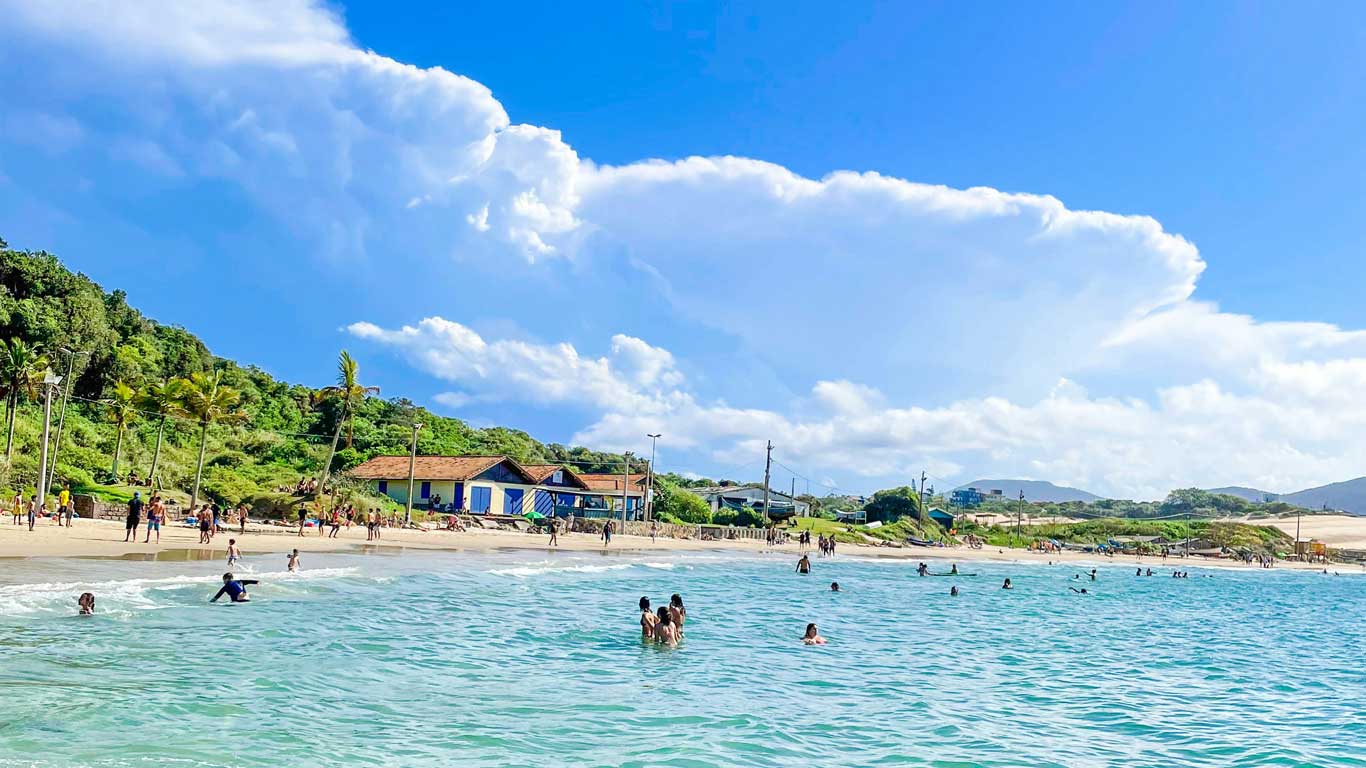 playa-ingleses-florianopolis Playa de aguas cristalinas y arena blanca en Florianópolis, conocida como Playa de los Ingleses. Varias personas disfrutan del mar y la orilla, mientras una zona verde con palmeras y casas tradicionales en el fondo complementa el paisaje. El cielo despejado con nubes esponjosas resalta el ambiente tropical y relajante de este destino.