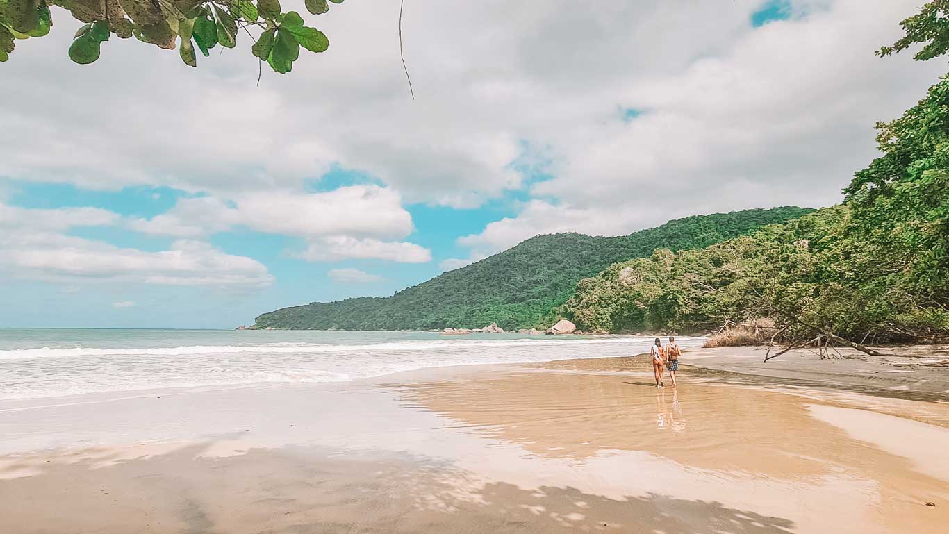 La imagen muestra una playa paradisíaca en Trindade, Río de Janeiro, con arena dorada, aguas cristalinas y un paisaje rodeado de montañas cubiertas de vegetación. Dos personas caminan por la orilla, dejando huellas en la arena mojada, mientras el cielo azul con algunas nubes crea un ambiente sereno. A la derecha, la selva tropical se extiende hasta la playa, resaltando la belleza natural y salvaje del lugar.