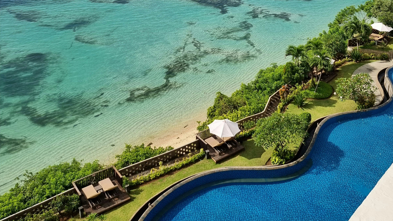 Una vista aérea de un lujoso resort en Bali, Indonesia, con una piscina infinita de azulejos azules que se funde visualmente con las aguas cristalinas del océano. La playa de arena blanca está rodeada de exuberante vegetación tropical, con tumbonas y sombrillas ofreciendo espacios de relajación. La combinación de colores vibrantes y el diseño elegante crean un ambiente de ensueño en este destino paradisíaco.