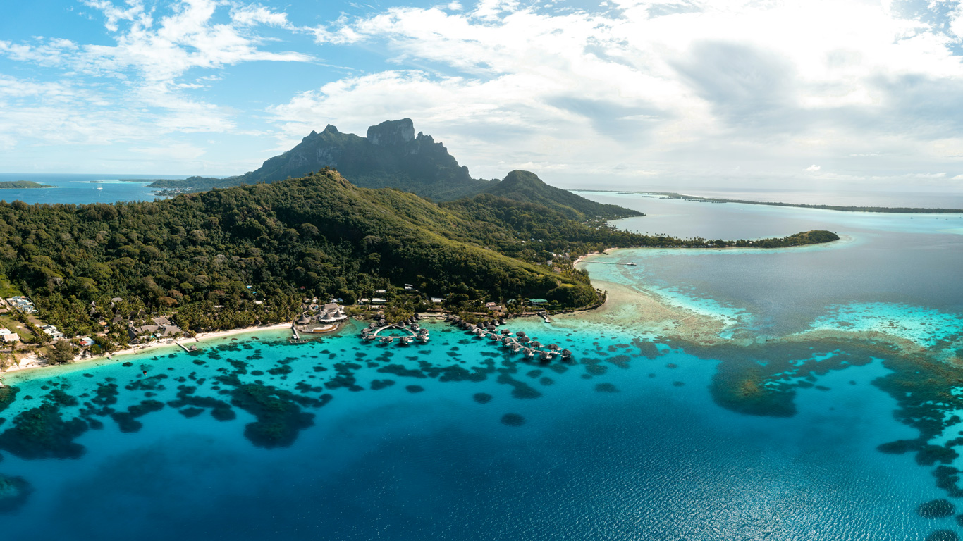 Una vista aérea espectacular de Bora Bora, Polinesia Francesa, con su icónica montaña rodeada de exuberante vegetación y lagunas turquesas. En el agua cristalina, se pueden ver bungalows flotantes conectados por pasarelas, ofreciendo un alojamiento de lujo. Los arrecifes de coral y las sombras en el mar agregan profundidad a esta imagen de ensueño en el paraíso tropical.