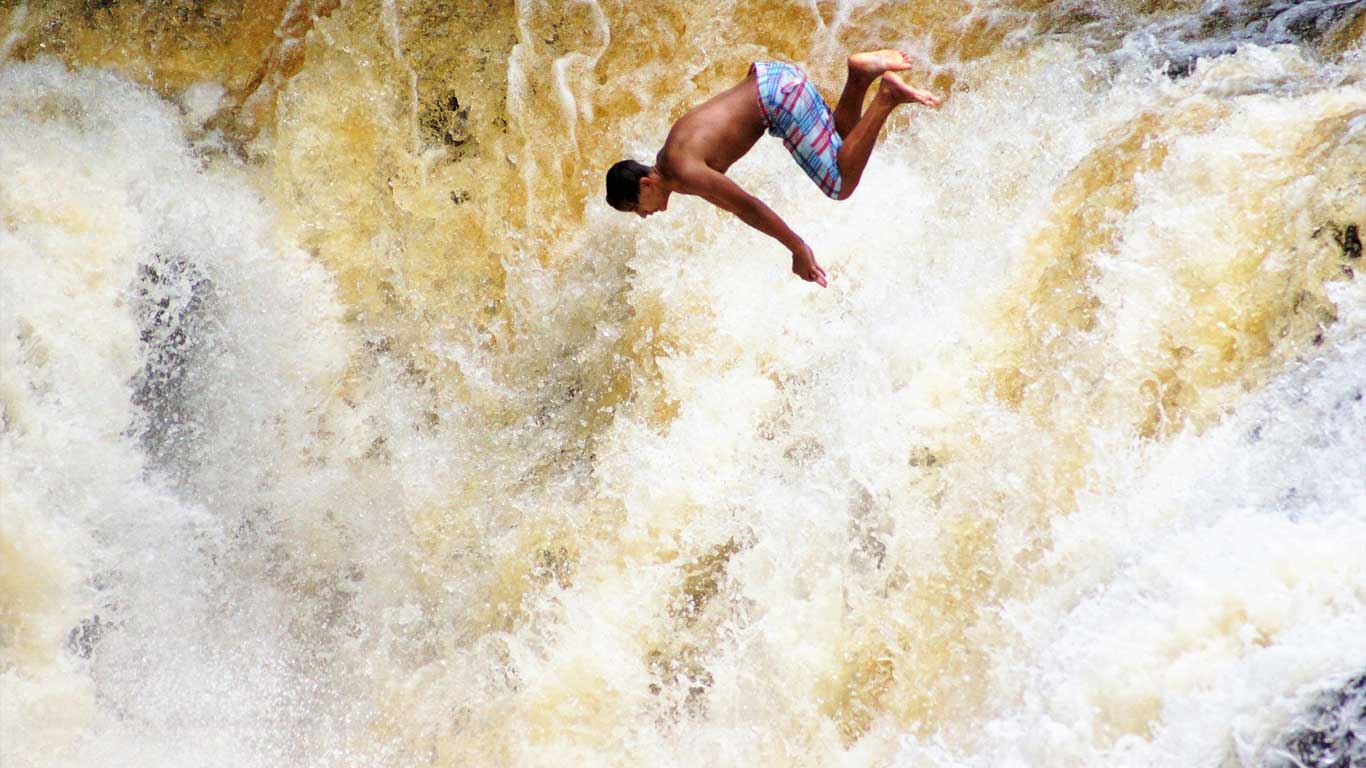 Un joven en traje de baño realiza un salto acrobático en medio de una cascada en Brotas, São Paulo. El agua cae con fuerza, creando un fondo dinámico de espuma y corriente turbulenta. La imagen captura un momento de adrenalina y aventura en este destino conocido por sus deportes extremos y ecoturismo.