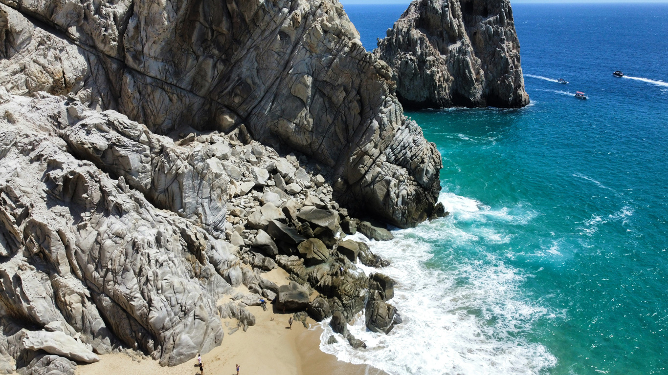 Una espectacular vista de una playa escondida en Cabo San Lucas, México, rodeada de imponentes formaciones rocosas. Las olas del océano turquesa chocan contra las piedras mientras algunas personas exploran la arena dorada. En el fondo, pequeñas embarcaciones navegan cerca de los icónicos acantilados, creando una escena salvaje y paradisíaca en la costa del Pacífico.