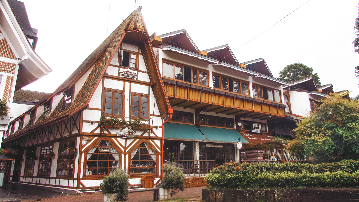 Edificio de estilo alpino con entramado de madera y techo inclinado en Campos do Jordão, São Paulo. La fachada blanca con detalles marrones y flores en las ventanas le da un encanto europeo, evocando una villa suiza. El lugar alberga tiendas y restaurantes, creando una atmósfera acogedora en este popular destino de montaña.