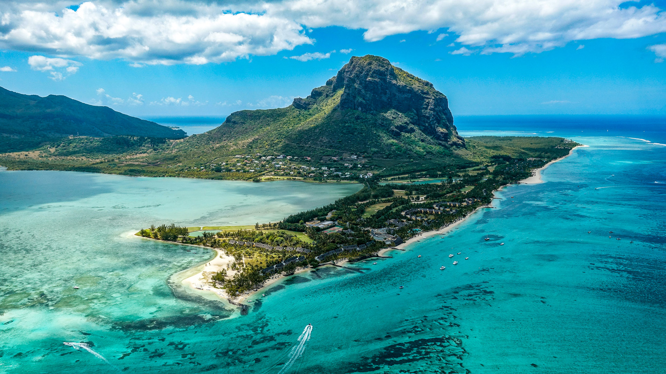 Una impresionante vista aérea de Le Morne Brabant en las Islas Mauricio, una montaña icónica rodeada por lagunas turquesas y playas de arena blanca. La exuberante vegetación cubre la isla, mientras pequeñas embarcaciones navegan por las aguas cristalinas. El paisaje combina montañas, mar y resorts de lujo, creando un destino tropical paradisíaco.