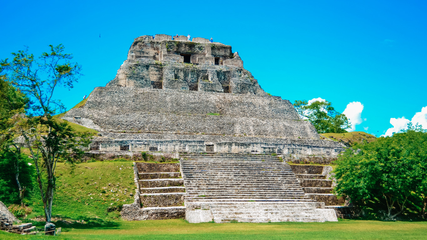 Una majestuosa pirámide maya de piedra, ubicada en medio de una exuberante vegetación bajo un cielo azul brillante. La estructura muestra su diseño escalonado con amplias escaleras que conducen a la cima, donde algunos visitantes disfrutan de la vista panorámica. Este sitio arqueológico representa la grandeza de la civilización maya y su impresionante legado arquitectónico.