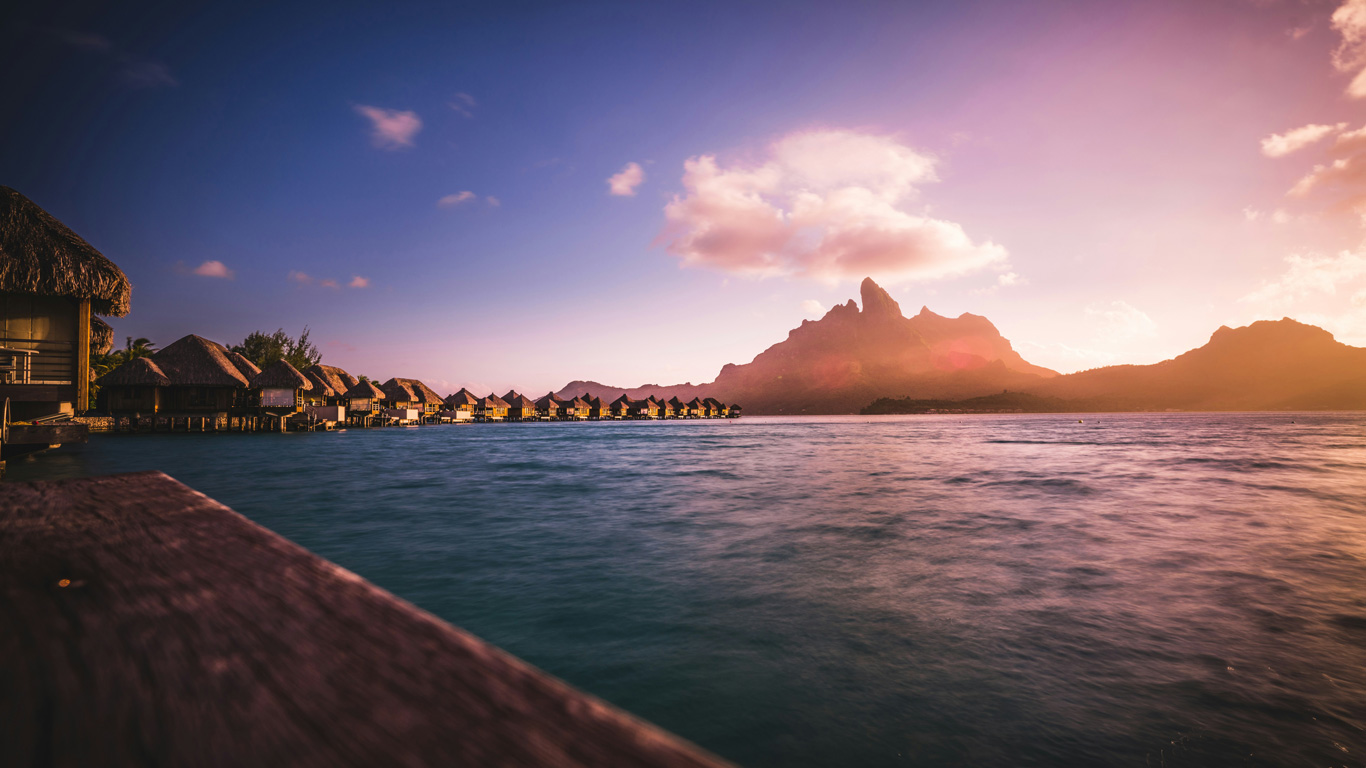 Un atardecer mágico en Bora Bora, Polinesia Francesa, con el sol tiñendo el cielo de tonos dorados y rosados sobre el icónico monte Otemanu. Bungalows flotantes con techos de paja se alinean sobre las tranquilas aguas de la laguna, reflejando la luz cálida del ocaso. La escena transmite lujo y tranquilidad en este destino tropical de ensueño.