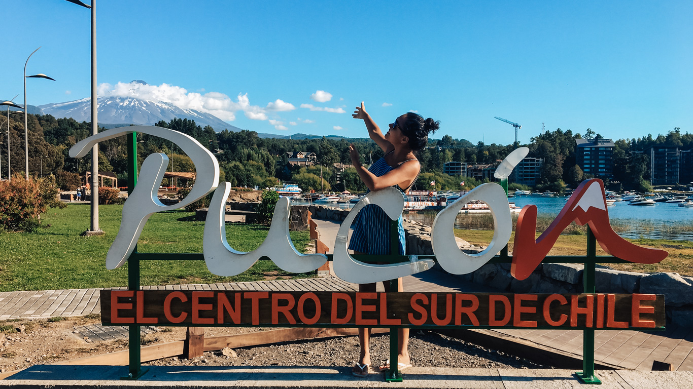 Una fotografía turística en Pucón, Chile, con una mujer posando junto al letrero colorido que dice "Pucón, el centro del sur de Chile". En el fondo, el imponente volcán Villarrica con su cumbre nevada domina el paisaje, rodeado de vegetación, un lago con embarcaciones y modernos edificios. El cielo despejado y el ambiente vibrante reflejan la belleza natural y el encanto de este destino chileno.