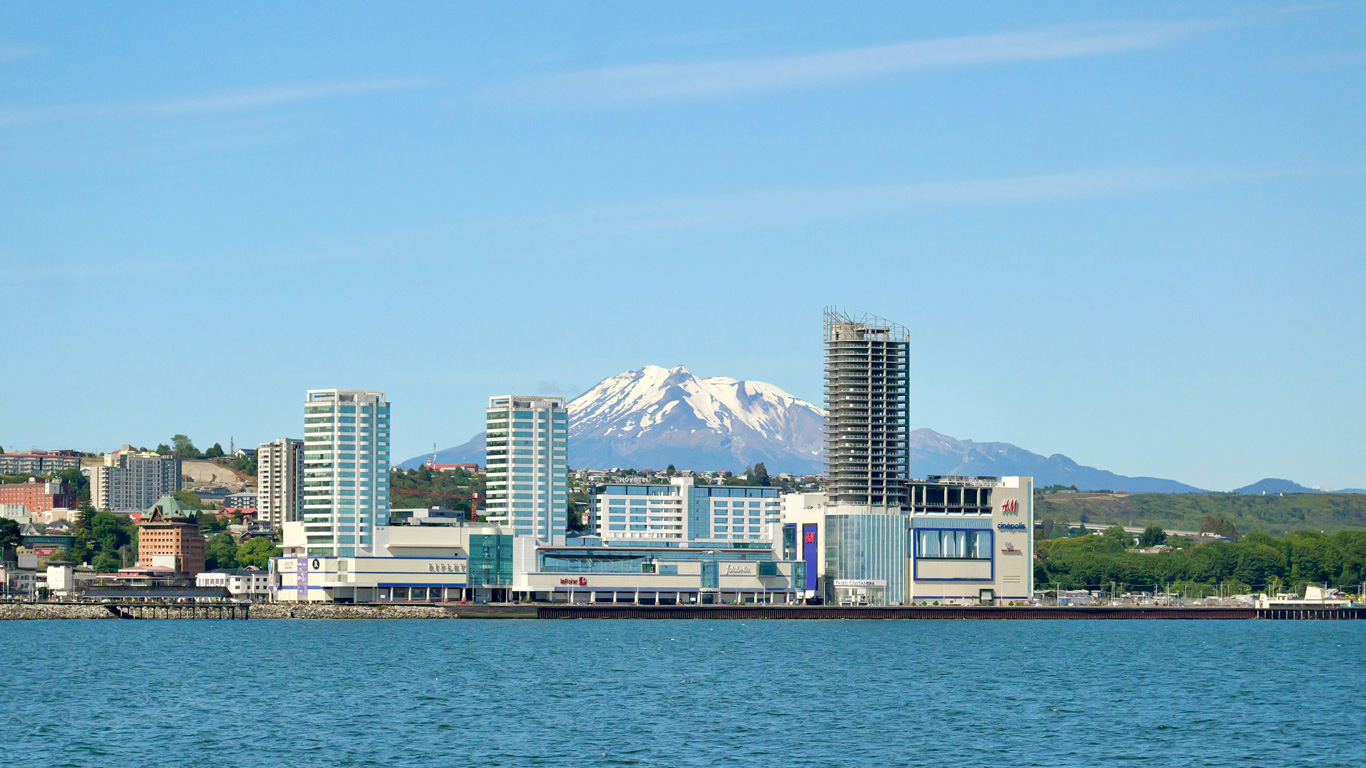 Vista panorámica de la ciudad de Puerto Montt, Chile, con modernos edificios frente al mar y el imponente volcán Calbuco nevado en el fondo. El agua azul del océano refleja el cielo despejado, mientras que la arquitectura urbana contrasta con el paisaje natural montañoso. La imagen resalta la combinación única de ciudad y naturaleza en este destino del sur de Chile.