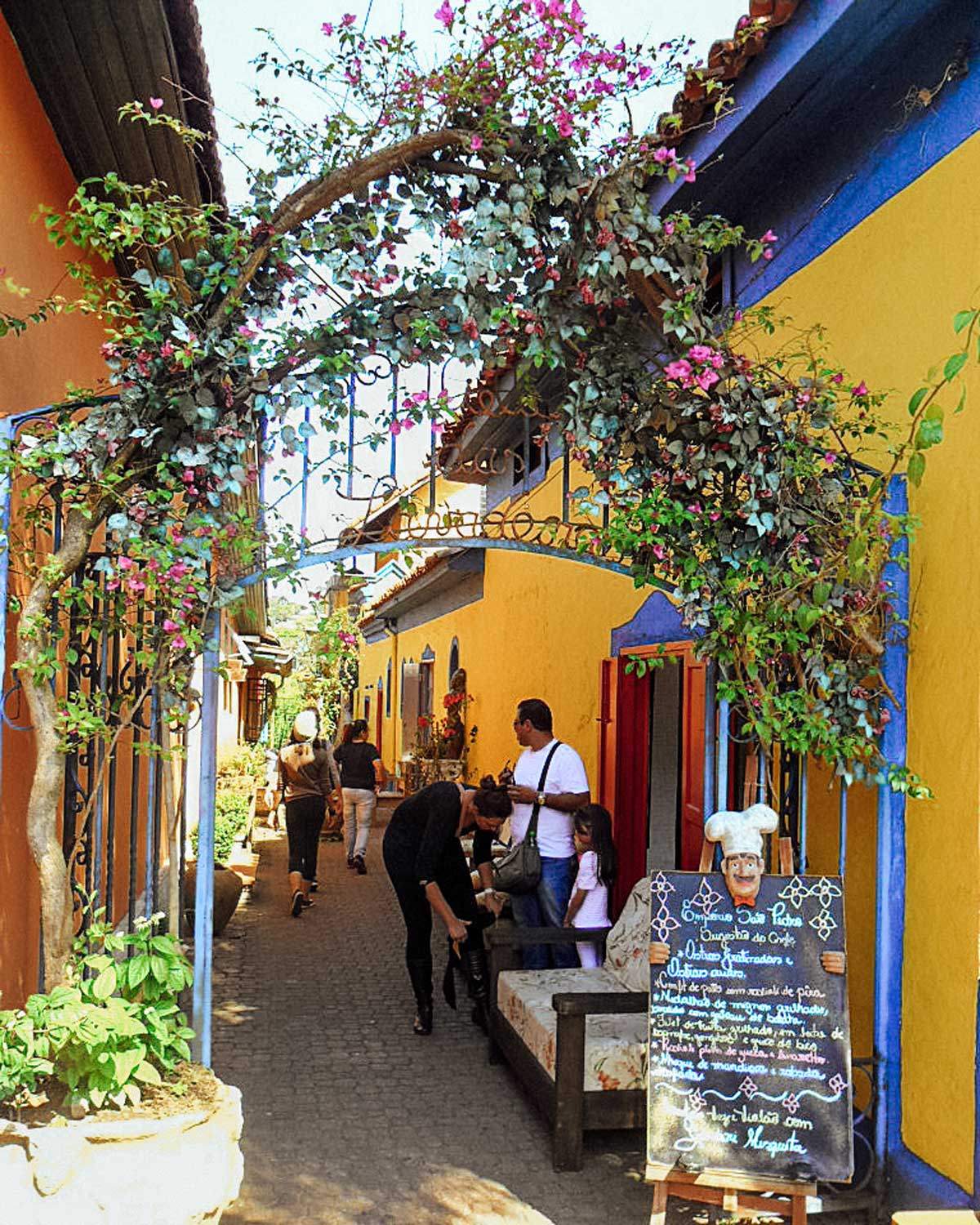 Un callejón pintoresco en Embu das Artes, São Paulo, con casas de colores vibrantes, en amarillo y azul, decoradas con flores y detalles rústicos. Un arco cubierto de enredaderas y flores rosadas enmarca la entrada, mientras varias personas pasean y exploran el lugar. En primer plano, un cartel con un muñeco de chef anuncia un café, resaltando el ambiente acogedor y artístico de esta encantadora ciudad.