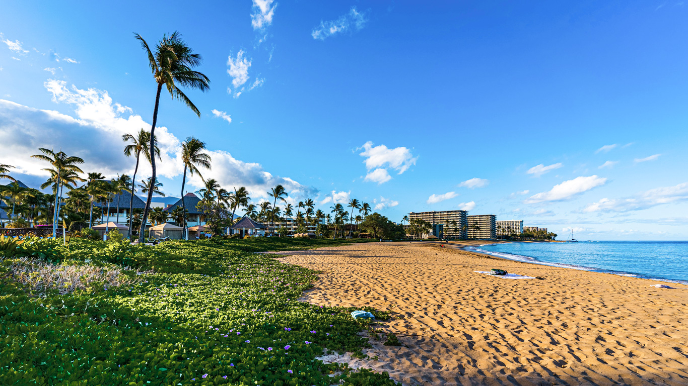 Una playa de arena dorada en la isla de Maui, Hawái, con aguas cristalinas y un cielo azul brillante con algunas nubes dispersas. Palmeras altas se mecen con el viento, rodeadas de vegetación tropical y lujosos resorts frente al mar. La escena transmite una atmósfera relajante y paradisíaca, ideal para unas vacaciones en el paraíso hawaiano.