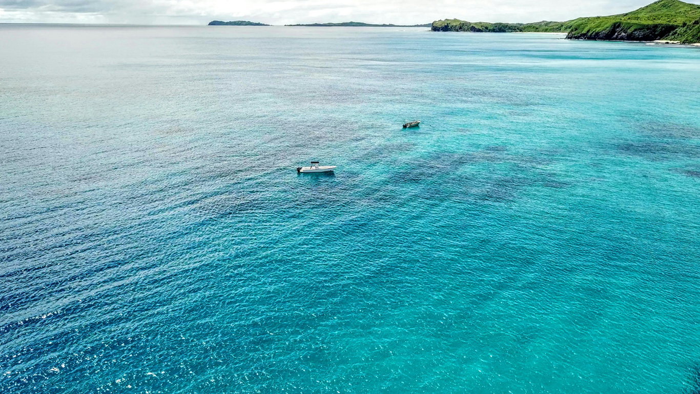 Una vista aérea del océano en las Islas Fiyi, con aguas cristalinas en tonos turquesa y azul profundo. Dos pequeñas embarcaciones flotan tranquilamente sobre la superficie, rodeadas por el mar infinito. En el horizonte, se pueden ver islas cubiertas de exuberante vegetación, creando una escena tropical y paradisíaca.