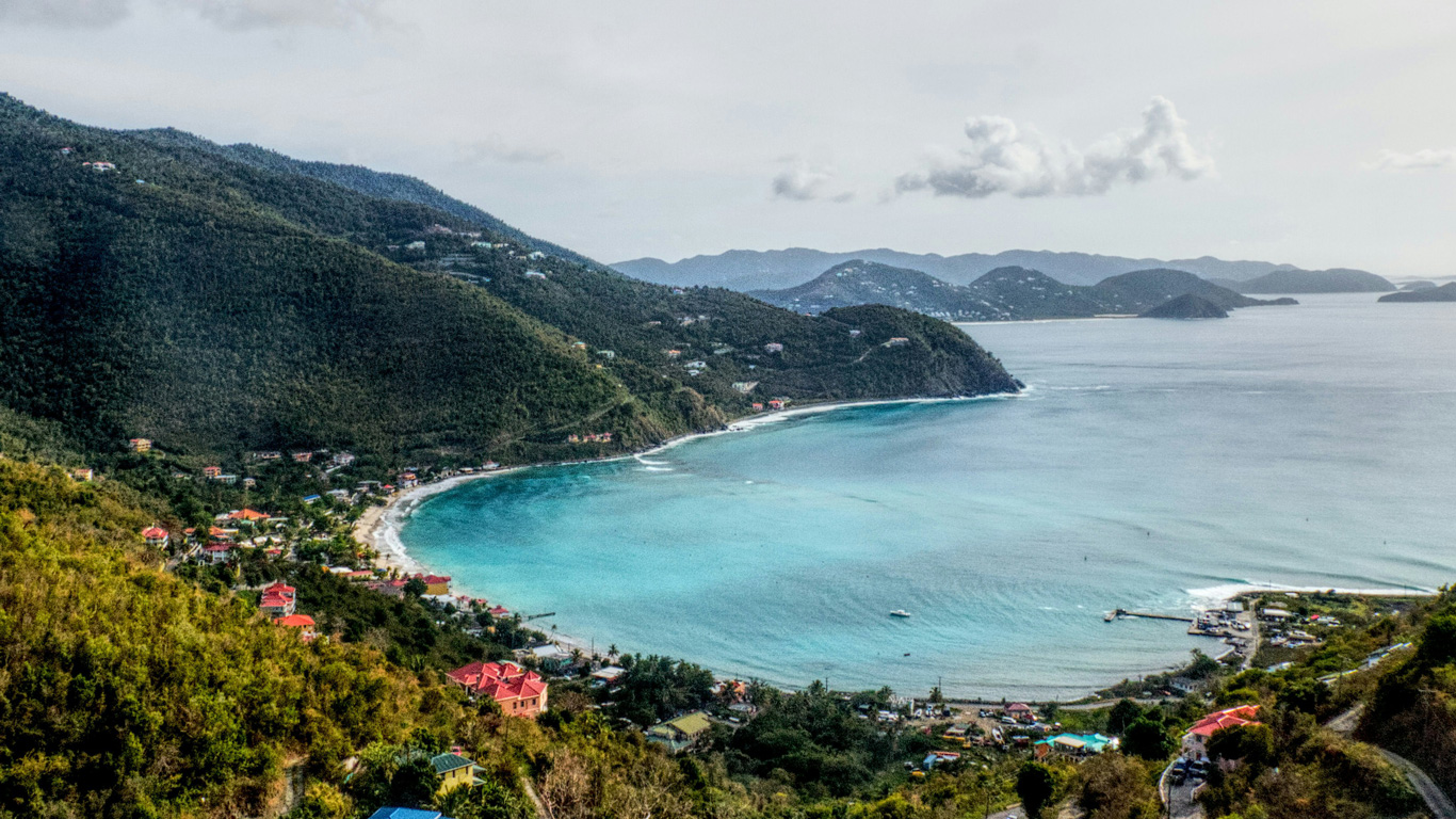 Una vista panorámica de una bahía en las Islas Vírgenes Británicas, con aguas turquesas y arenas blancas rodeadas de colinas verdes cubiertas de vegetación. Pequeñas casas y edificios con techos rojos se esparcen a lo largo de la costa y las laderas, mientras algunas embarcaciones flotan en el mar tranquilo. En el fondo, otras islas montañosas emergen en el horizonte bajo un cielo parcialmente nublado.