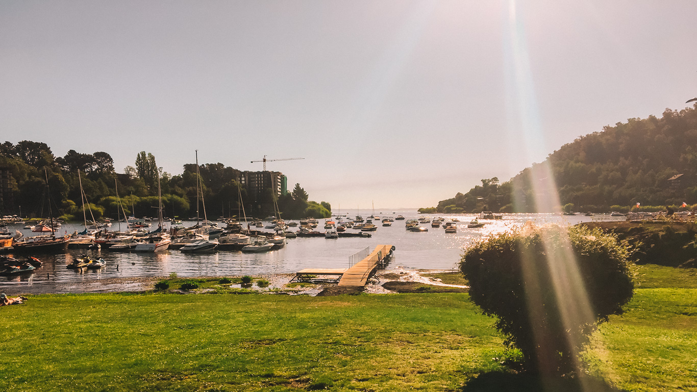 Un atardecer en el lago Villarrica, en Pucón, Chile, con el sol reflejándose sobre el agua y creando un efecto dorado en el paisaje. En el muelle, decenas de embarcaciones y veleros están anclados, rodeados de colinas verdes y edificios modernos en la distancia. El césped vibrante en primer plano y los rayos de sol añaden un toque cálido y sereno a esta escena natural.