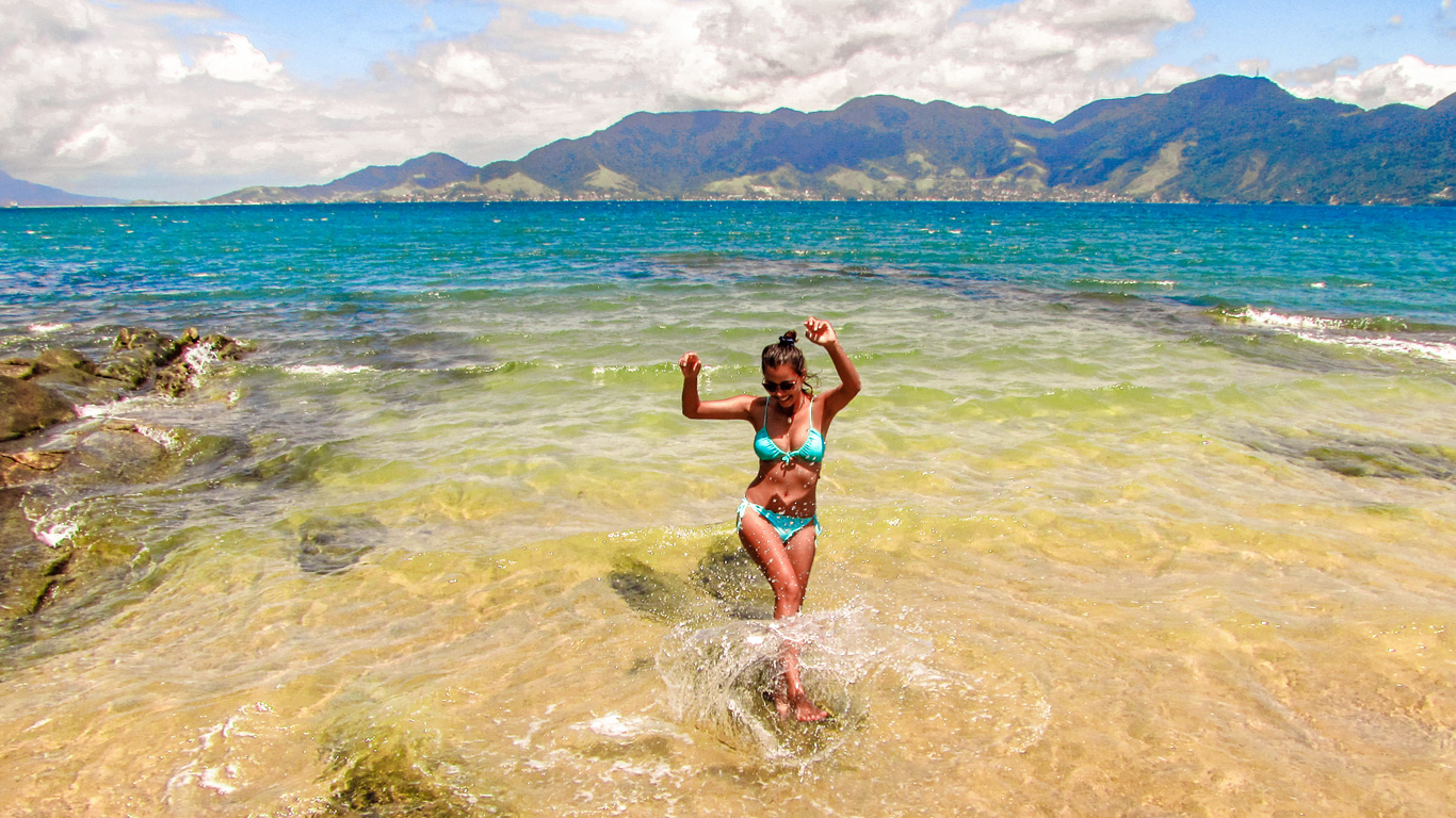 Una mujer con bikini azul turquesa disfruta del mar cristalino en una playa tropical, levantando los brazos con alegría mientras el agua salpica a su alrededor. Detrás de ella, el océano azul profundo se extiende hasta el horizonte, con montañas verdes en la distancia y un cielo parcialmente nublado. La imagen captura un momento de felicidad y conexión con la naturaleza en un destino paradisíaco.