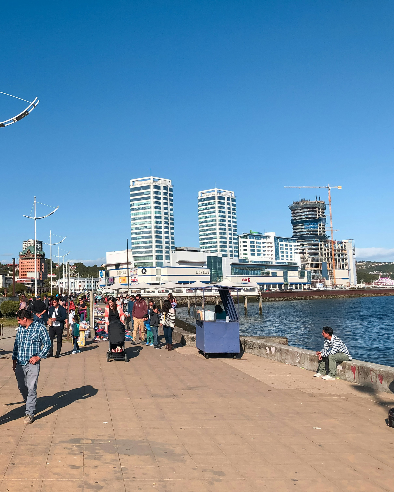 Un animado paseo costero en Puerto Montt, Chile, con personas caminando, vendedores ambulantes y un hombre sentado en el borde del malecón mirando el mar. Al fondo, los modernos edificios del centro comercial y hoteles se elevan bajo un cielo azul despejado. La imagen refleja el contraste entre la vida urbana y el paisaje marítimo de esta ciudad portuaria del sur de Chile.