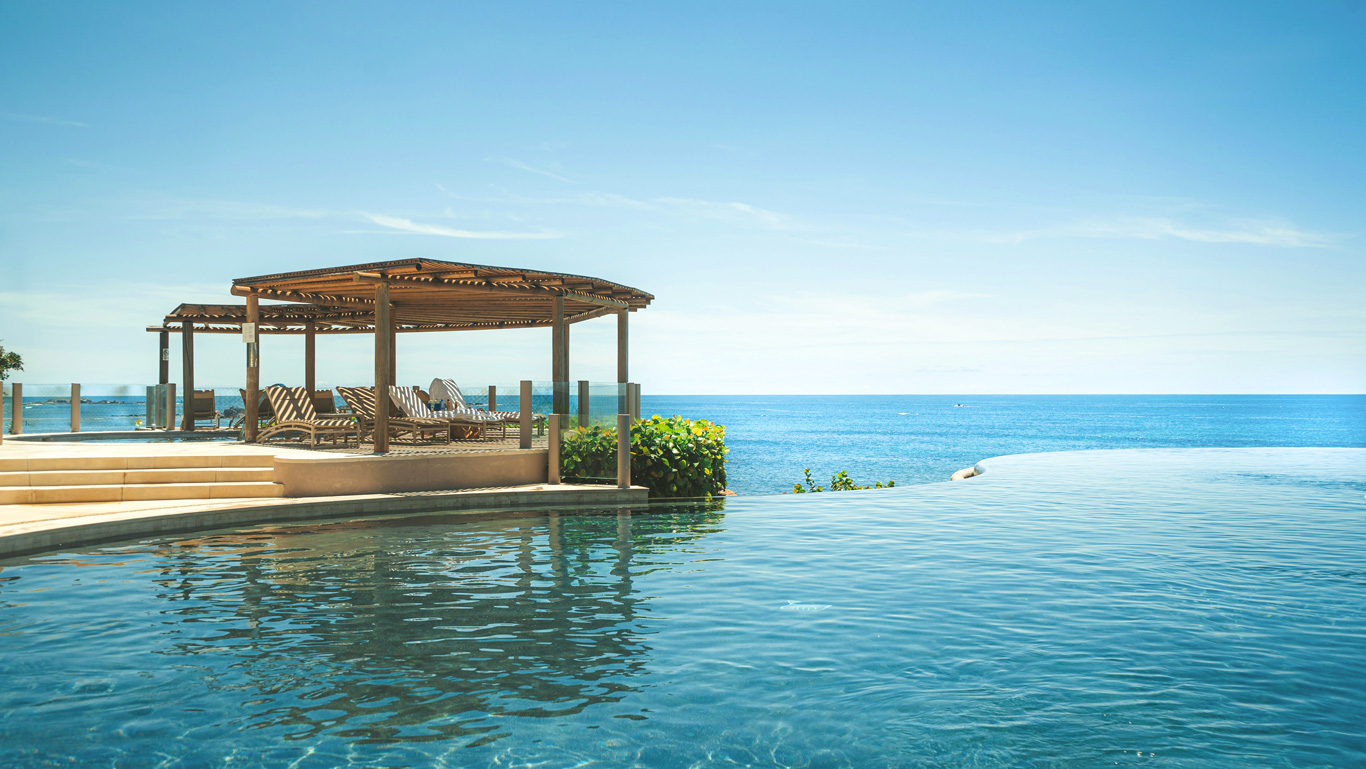 Una lujosa piscina infinita con agua cristalina se fusiona visualmente con el océano azul en el horizonte. A un lado, una pérgola de madera con tumbonas ofrece un espacio sombreado para el descanso, rodeado de un paisaje tropical. El cielo despejado y el ambiente sereno evocan una sensación de tranquilidad y exclusividad en este destino de playa.